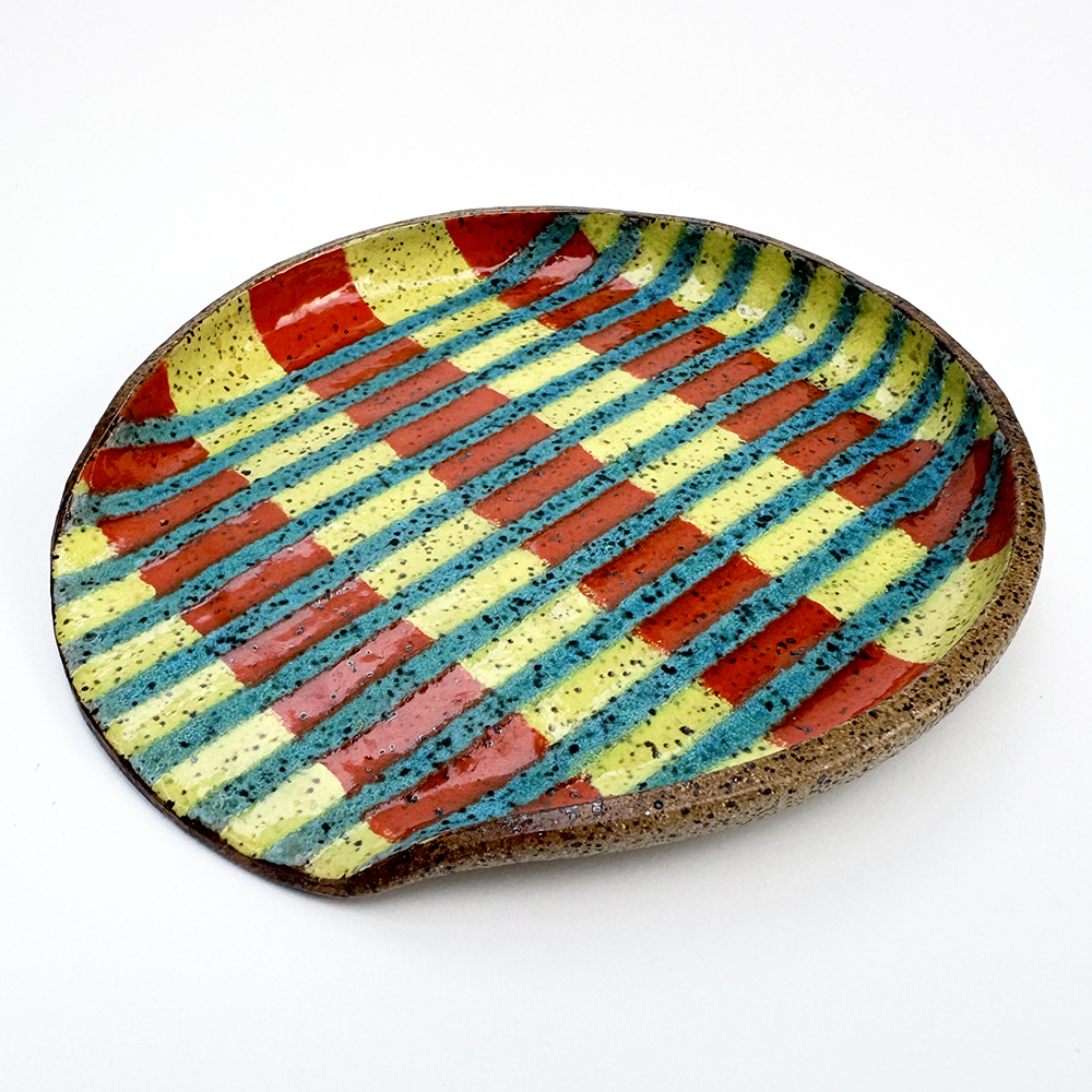 A speckled spoonrest features thin matte turqouise lines that run vertically across the piece. Behind them, thick glossy yellow and orange stripes make up the background.