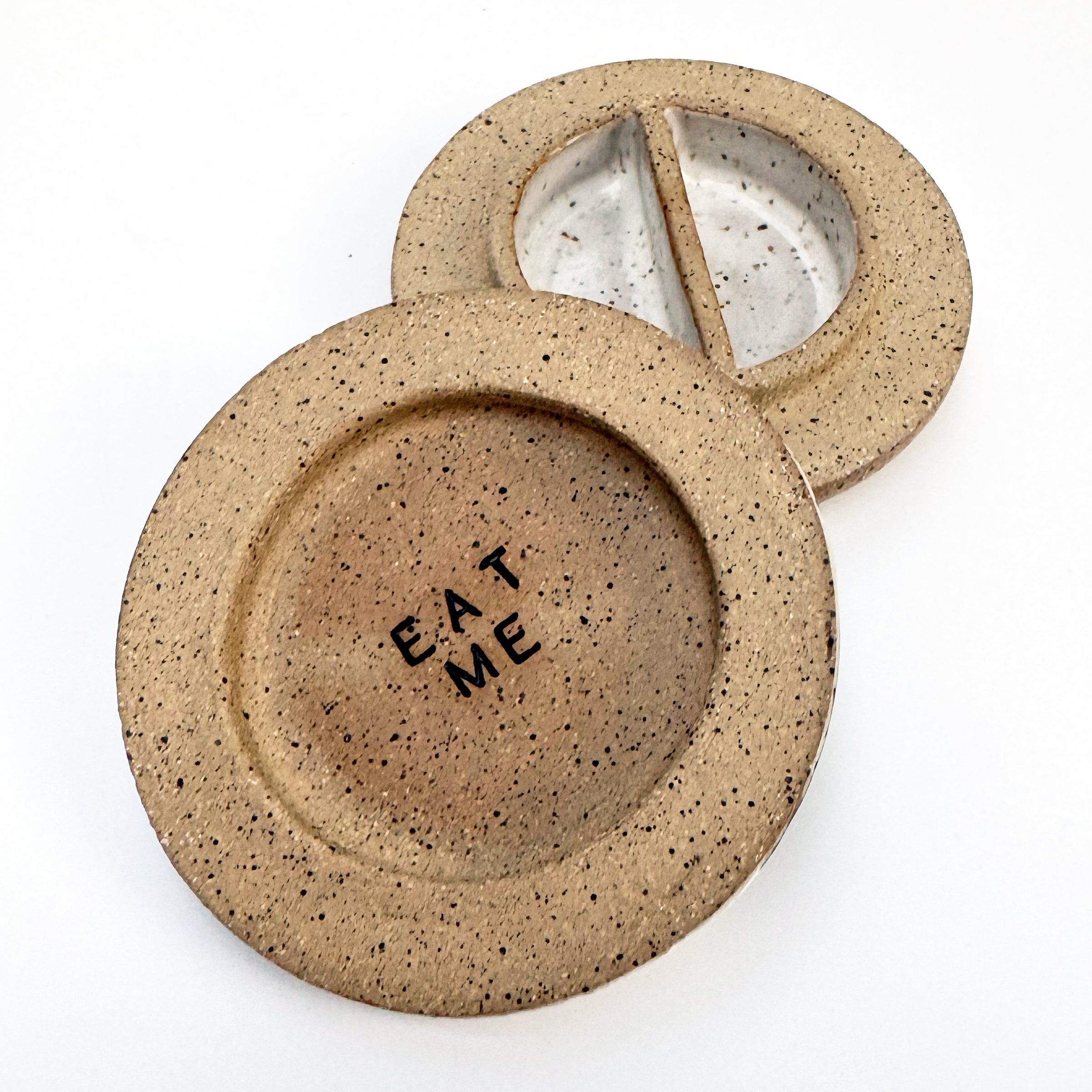 The interior is divided into two-compartments that are glazed white, and the black speckles in the clay show through the glaze. The underside of the lid says 'eat me' in black glaze inlaid into stamped letters.