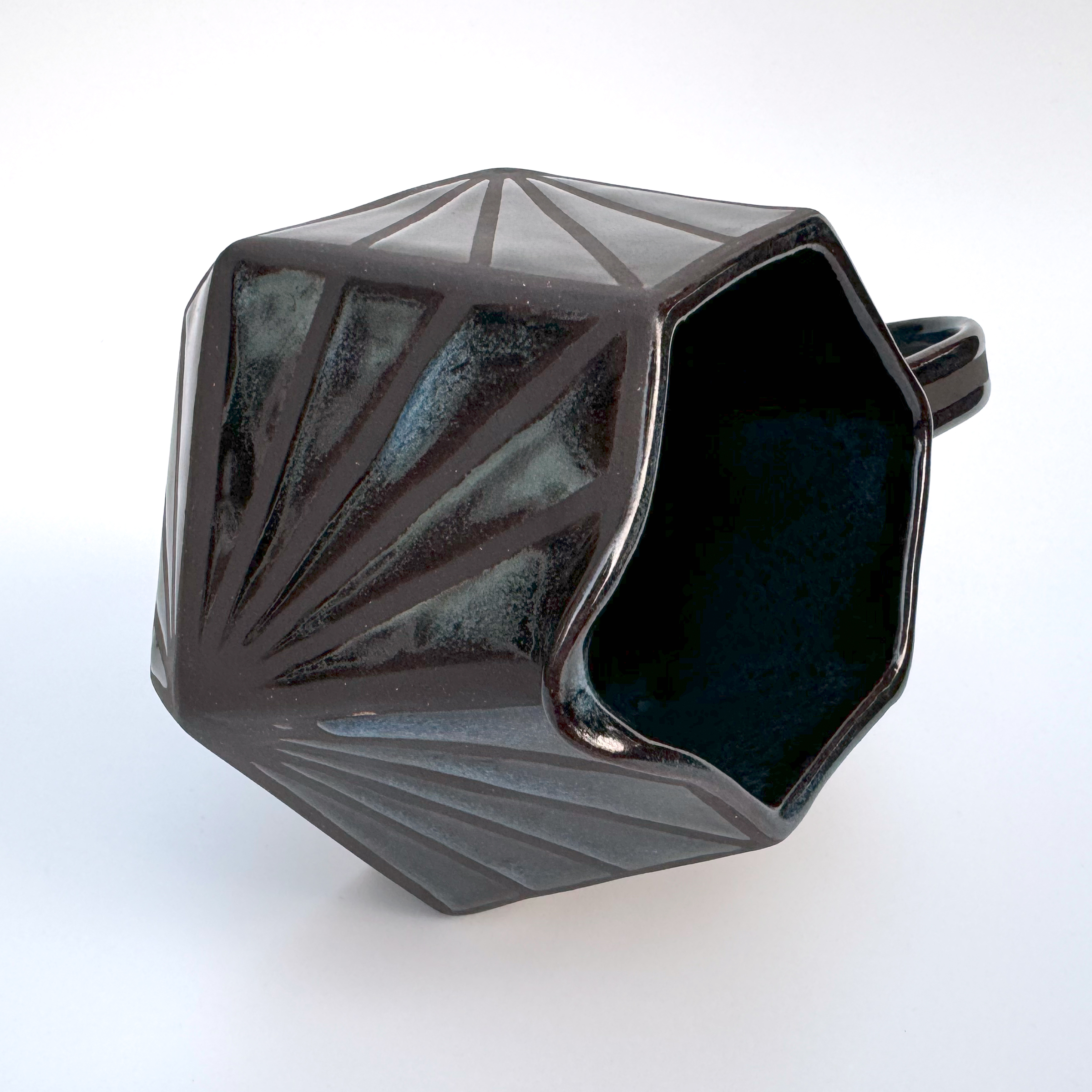 This black stoneware pitcher has a squat, geometric form with a hexagonal base and opening. The form flares out towards the bottom and each of the six sides is made of two trapezoids - a larger one on top and a smaller on the bottom.