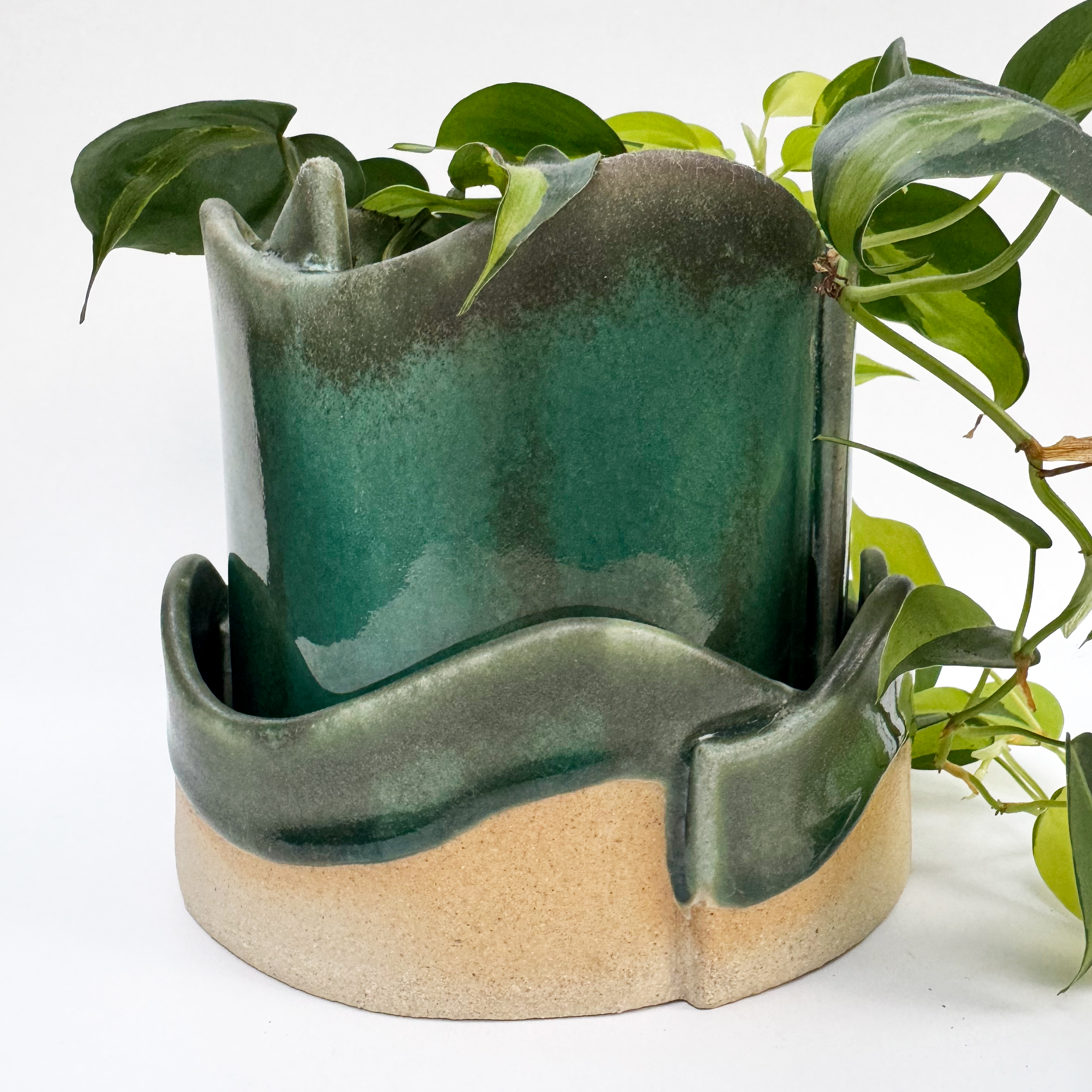 The planter and its drip tray both have wavy edges and sections of each overlap. The planter is glazed in a glossy teal green. Near the rim, the glaze is more matte and grey, and in the body, it is the bright crackled blue-ish green.