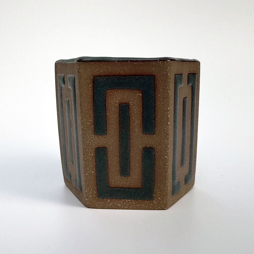 Hexagonal teacups in a medium brown stoneware have a maze-like pattern made of denim glaze on each of their sides.