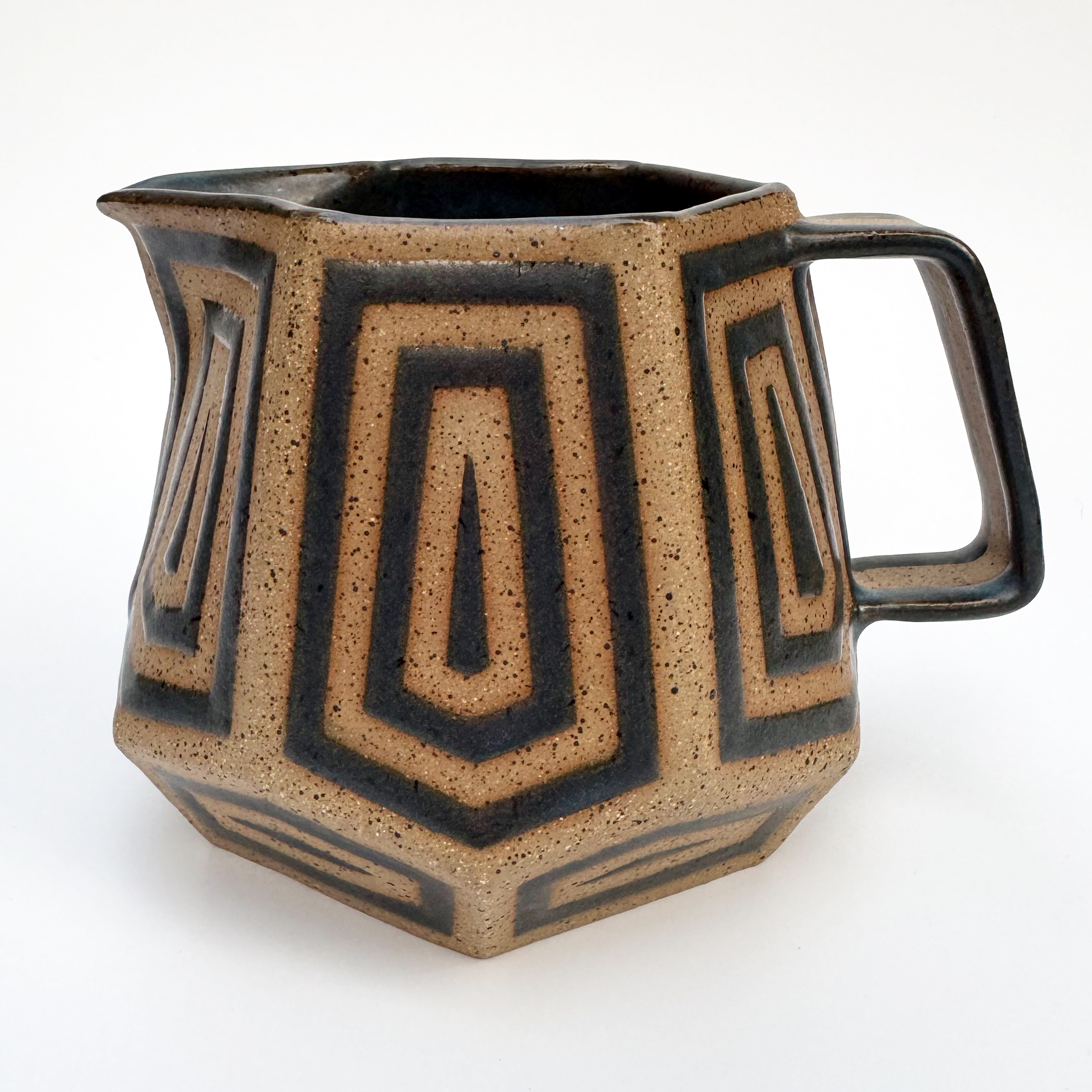 This tan speckled stoneware pitcher takes the form of a truncated hexagonal trapezohedron. The base and opening are hexagons, and there are 12 staggered pentagons around the sides. The upper ones are long and the bottom ones narrow.