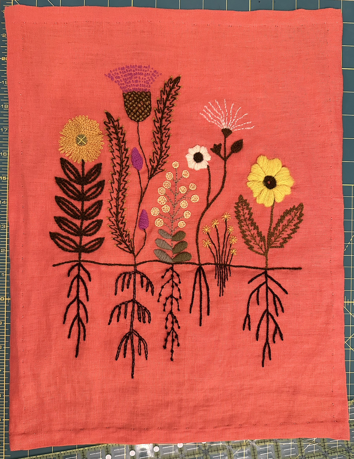 A variety of flowers and their roots are embroidered on a bright orange linen evenweave background in wool crewel yarn. The blooms are in a variety of stitch designs in shades of green, shades of yellow, a light peach, and also a bright pink. The roots are stitched in dark brown below a ground line.