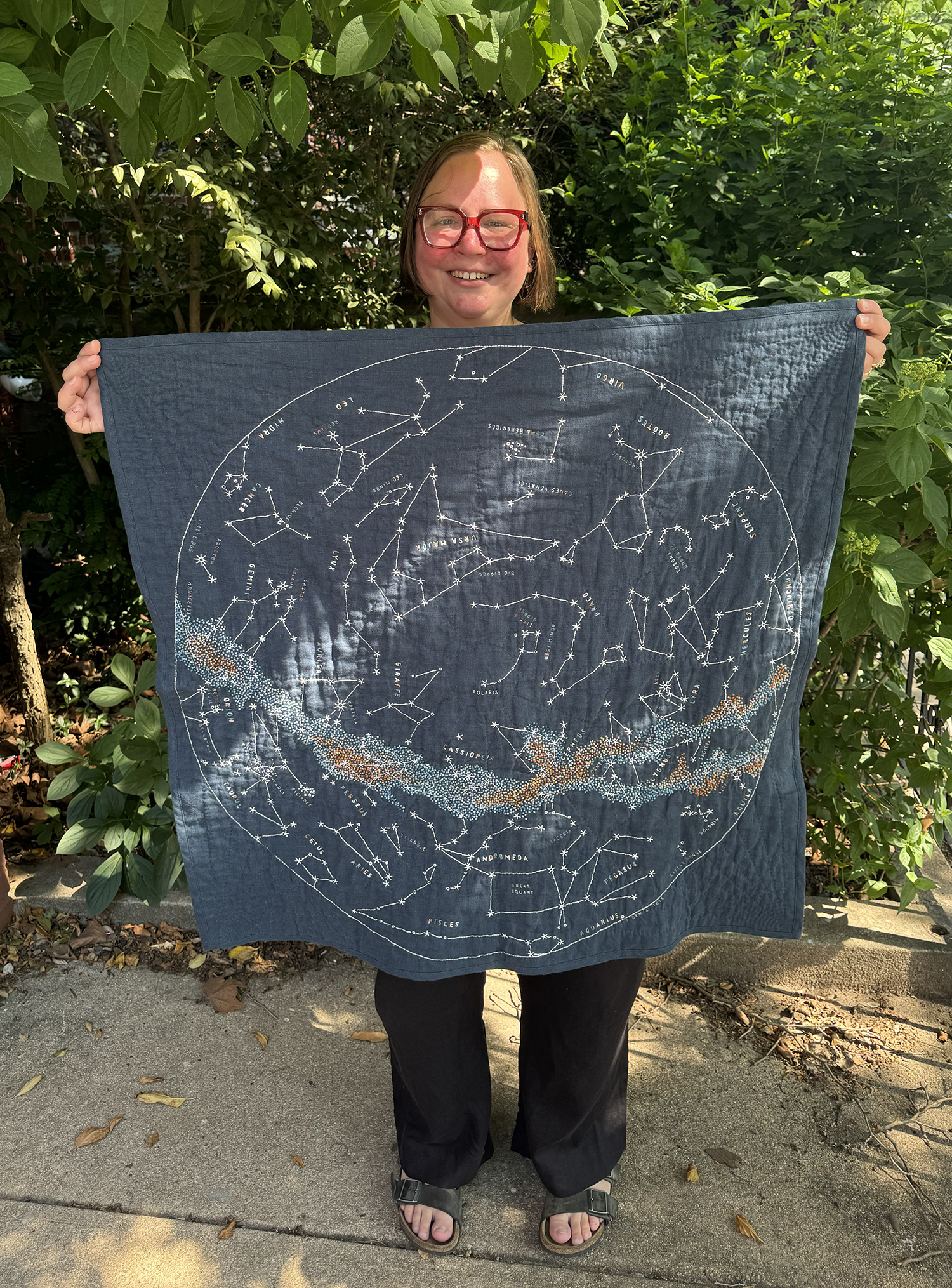 Me holding up my Northern Sky quilt. The fabric is a deep inky teal blue linen, the constellations of the Northern Hemisphere are outlined in white thread, and the Milky Way is a gradient of French knots in coppers, blues, greens, and greys. Constellation names are done in a gradient of the same colors as the Milky Way.