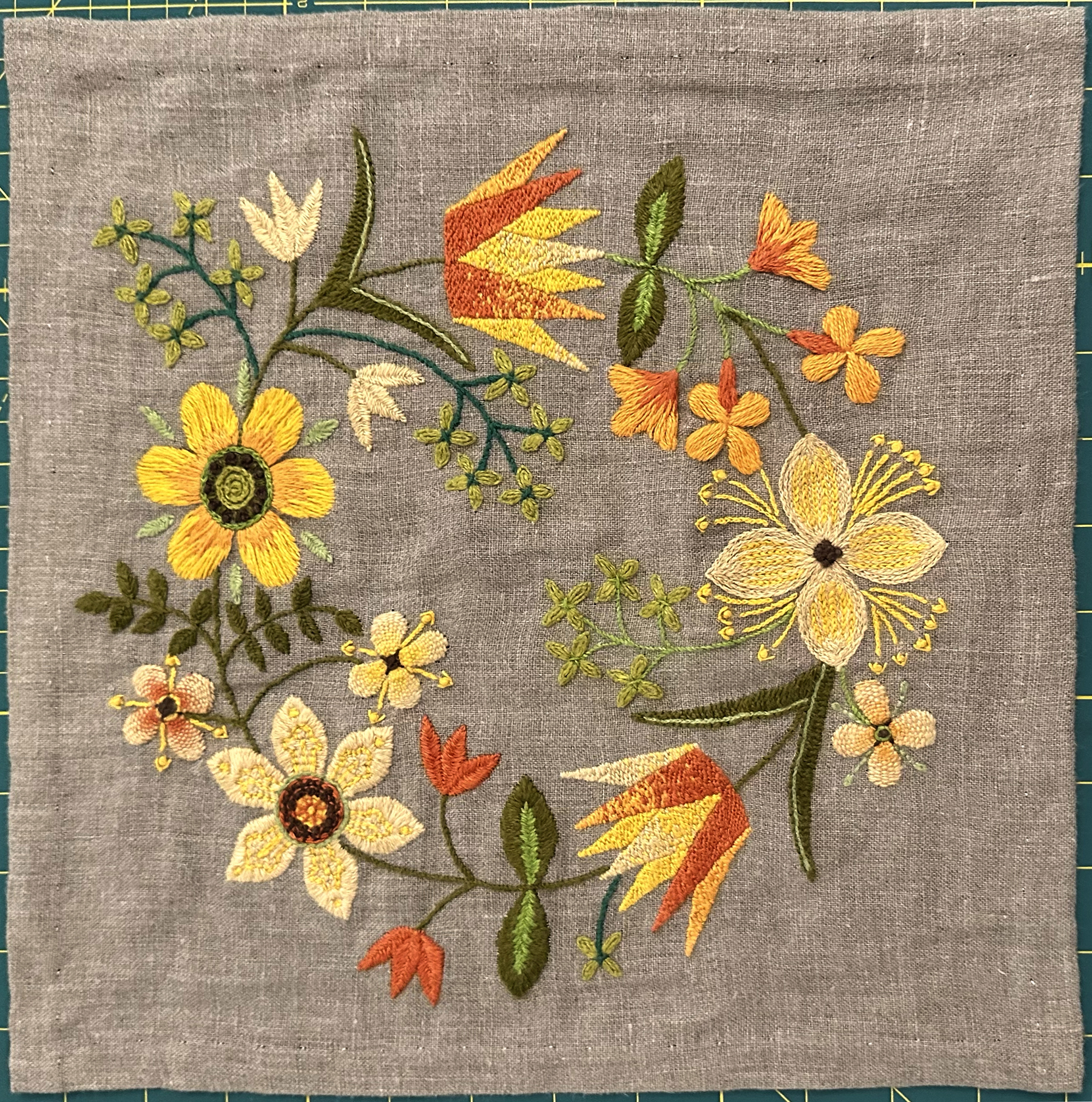 An embroidered wreath of a variety of flowers on a natural linen background. The stems are varying shades of greens, and the flowers are stitched in a pale peach, sunflower yellow, bright orange, firey red orange, and dark brown.