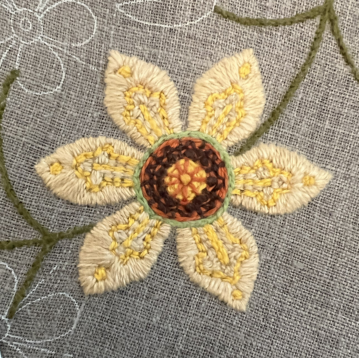 Another folk art-inspired flower: the center has a wheel-inspired design. The very center is yellow backstitch fill in a circular shape with a red orange wheel stitched on top of it, and just outside that there is a layer of red orange with dark brown stitched on top of it. The petals are a light peach satin stitch with yellow stitching on top in a pattern.