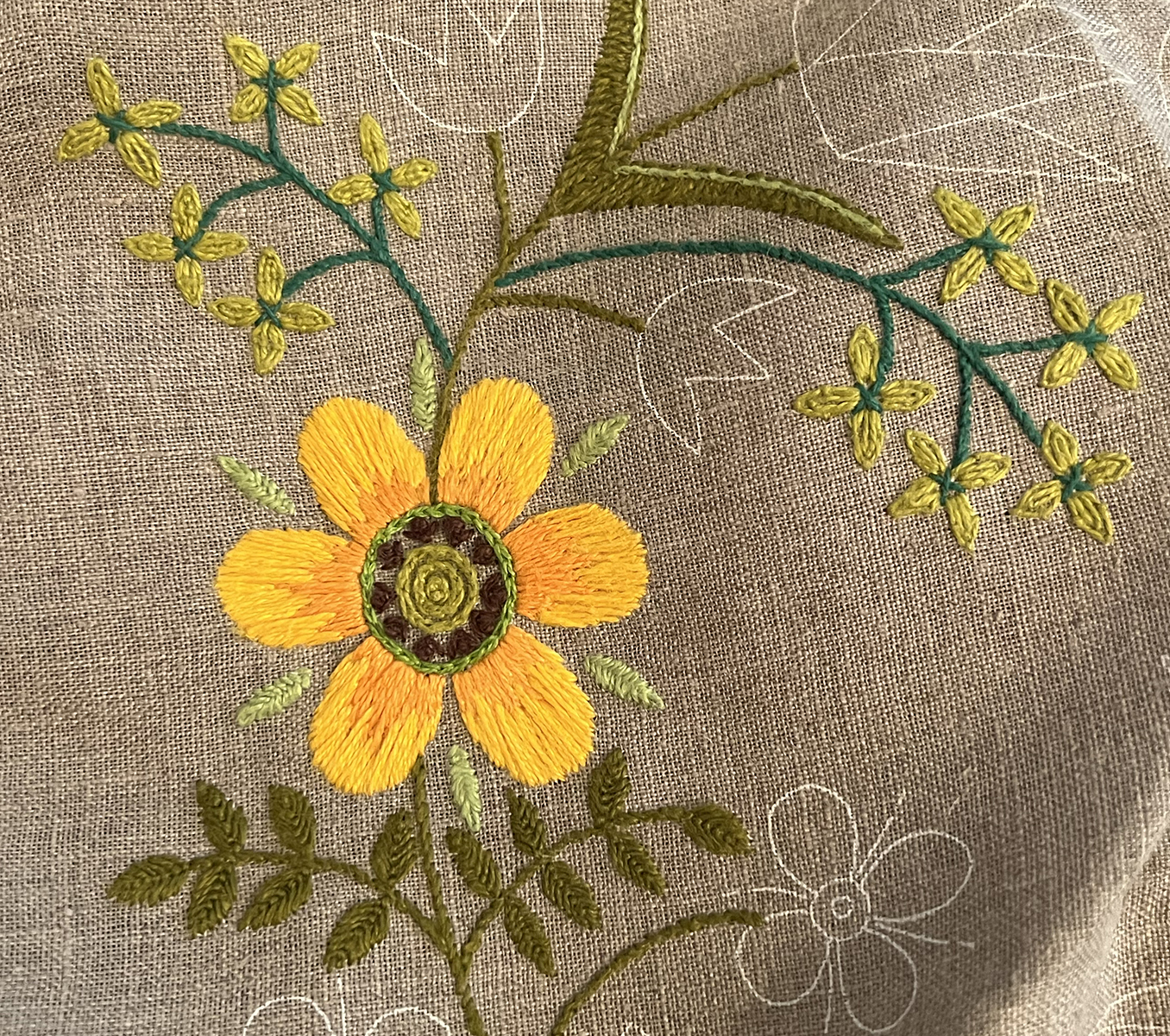 A six petaled flower's round petals are no longer in alternating yellow and yellow orange like the original pattern, but are threadpainted in long and shorts with the petals being more orange towards the center. The center of the flower references folk art stitches with varying greens in chain stitch and a layer of dark brown fly stitches with French knots in the center. Small light green clusters of leaves are done in closed fly stitch.