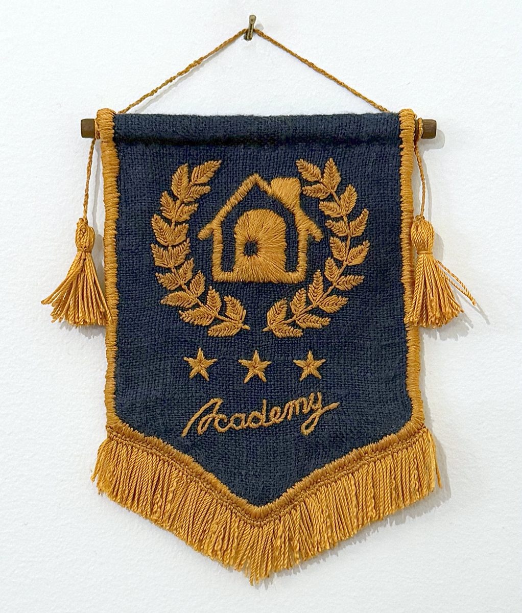 Happy Home Academy mini-pennant hanging on a white wall: the base is a deep slightly-teal navy linen with embroidered with gold silk thread. The pennant is vertically oriented and appears to be a rectangle that is slightly longer than it is wide with a short isocoles triangle right below it whose longest side is the bottom side of the rectangle. The design is embroidered in gold silk thread: There's a small house surrounded by a laurel wreath, three 5-pointed stars below it, and the word 'Academy' below the stars. The pennant's side and bottom edges are wrapped in gold silk thread, and there's a fringe hanging from the bottom. The pennant is hung with gold silk thread attached to a walnut-toned wooden dowel that runs through the top of the pennant, and a gold tassle hangs from each end of the wooden rod.