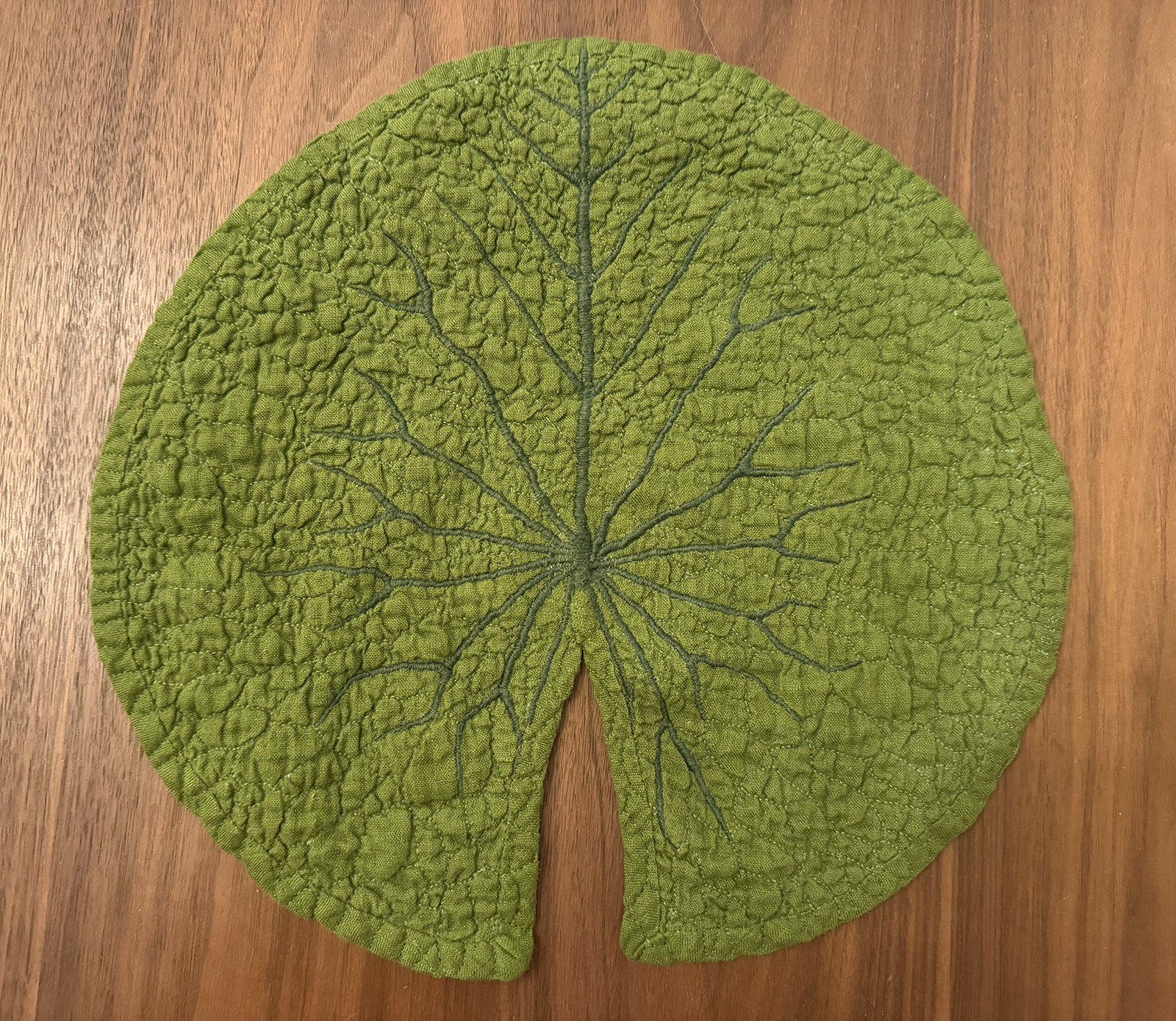 An olive green linen hand quilted, hand embroidered lilypad: the main veins are embroidered in a darker green satin stitch and other veins are quilted in an olive thread that nearly matches the olive green linen fabric.