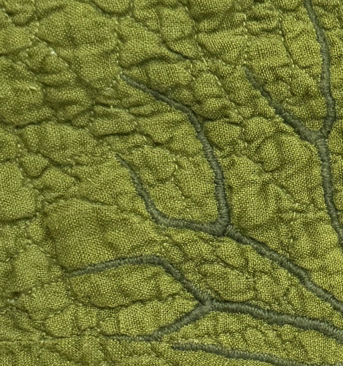 The darker green satin stitches dome slightly to create a texture above the quilt, while the smaller veins recess into the quilted sections.