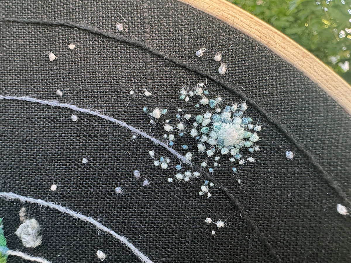 An embroidered supernova exploding in the Outer Wild solar system: it's a bunch of different French knots in various blues, teals, and whites on a black linen evenweave background. The center of the supernova is a large white French knot immediately bordered by slightly smaller aquamarine French knots. Other French knots in varying blues and whites spread out from that center. A tiny embroidered Attlerock is closer into the sun than this supernova.