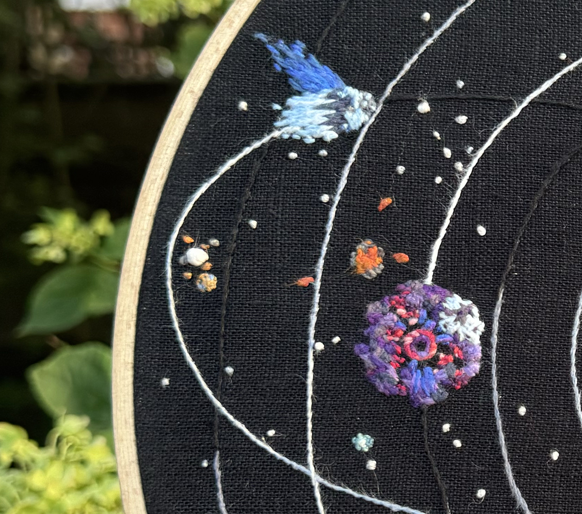 Brittle Hollow, Hollow's Lantern, the Interloper, the white hole, and White Hole Station stitched on a black linen background: Brittle Hollow primarily features short stitches in pinks, purples, blues, and white. The cracking surface of the planet is made up of hexagons in erraticly oriented satin stitch in bright purples, deep purples, navy, and some nearly grey toned purples and blues, too. Beneath them, blue crystaline shards in long straight stitches point towards the center, and small clusters of stitches in varying shades of pinks make up some of the insides of the planet. The very center is done in small black stitches in a spiral pattern with stem stitch in shades of purple encircling it which is in turn encircled by a circle of stem stitches in shades of pink. The upper right section of the planet encased in ice is represented by short stitches in varying shades of white and very light aqua with a deep purple crack stitched across it. Hollow's Lantern's volcanoes are done in small stitches of medium greys, and the lava is done in three shades of orange. Three lava balls are each made of a short stitch in a bright orange that is split by another short stitch in a redder orange. The Interloper's rocky surface is made in short back stitches of grey tones with white and light aqua stitches on its snowy surface. The two tails of the comet are in gradients stitched in long and shorts of varying blues. The white hole is a very large colonial knot of three strands of white linen thread, and very small French knots in grey and burnt orange are nearby to represent the comets and debris that came through the white hole. White Hole Station is made stitched in fine linen threads in two coppery gold tones on top of swoosh in stem stitch with pale blue thread to represent the ice. A very small supernova of just a single white French knot surrounded by a ring of smaller French knots in finer teal and aqua threads is nearby Brittle Hollow.