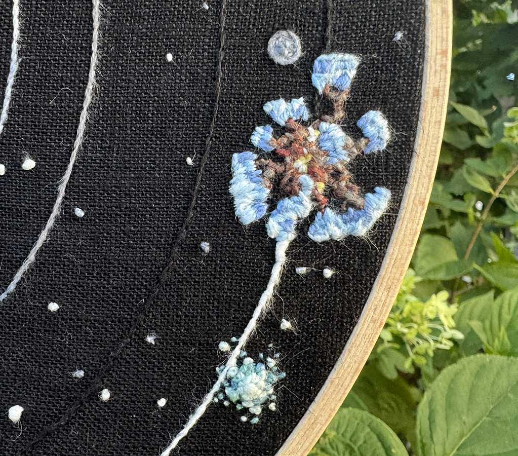 Dark Bramble and the Quantum Moon embroidered on a black linen background: Dark Bramble's spindly wooden body is stitched in what looks like a stem stitch in multiple colors. Each branch is a fake stem stitch in two different shades of browns, some more red, others more grey, and small thorns extend from the body and larger branches as well. There are small pockets of light coming from the bulbous center that are represented by light yellow and white short stitch clusters. The borken icy surface attached to the end of the branches is represented through satin stitches in varying shades of light blue; the surface sections are lighter blues than the underside fragments. The Quantum Moon is orbiting Dark Bramble in my piece, and it's made up of a blend of multiple grey fine linen threads stitched over each other, giving it a fuzzier quality than the Attlerock. An embroidered star going supernova is nearby: it's made of French knots in thin threads in shades of blues, teals, and white, and its center is a French knot in a thicker white surrounded tightly by French Knots in a thicker aquamarine.