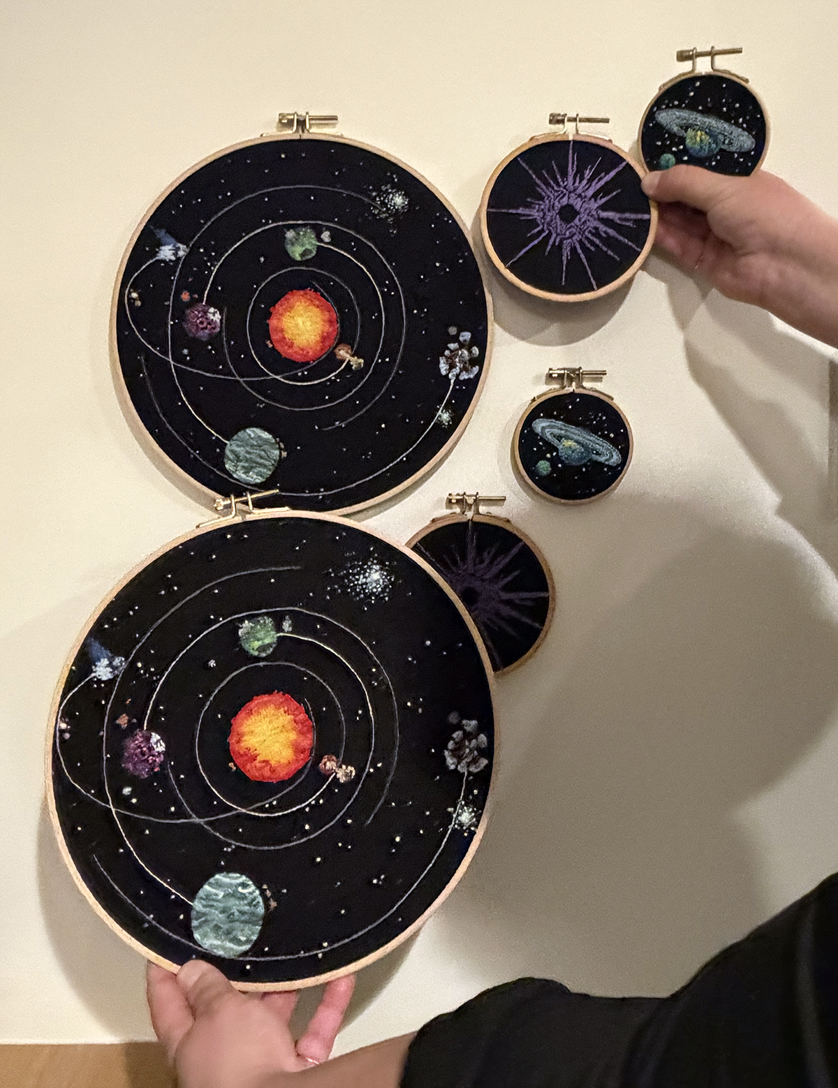 My hand is holding up the second edition of the three Outer Wilds embroidered hoops next to the original set hanging on my wall. They have subtle differences, as is expected with hand embroidered objects.