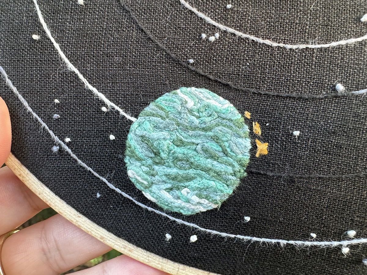 Giant's Deep and the probe launcher embroidered on a black linen background: Giant's Deep is embroidered in overlapping swirls of tight chain stitch in shades of teal and green with white swirls near the poles. Some of the swirls are just wavy, while others end in loops to evoke the cyclones present on the planet. The probe launcher is stitches in two shades of coppery golds in thin criss-crossed stitches, and it is split in three pieces. Stars and orbits can be seen in the background.