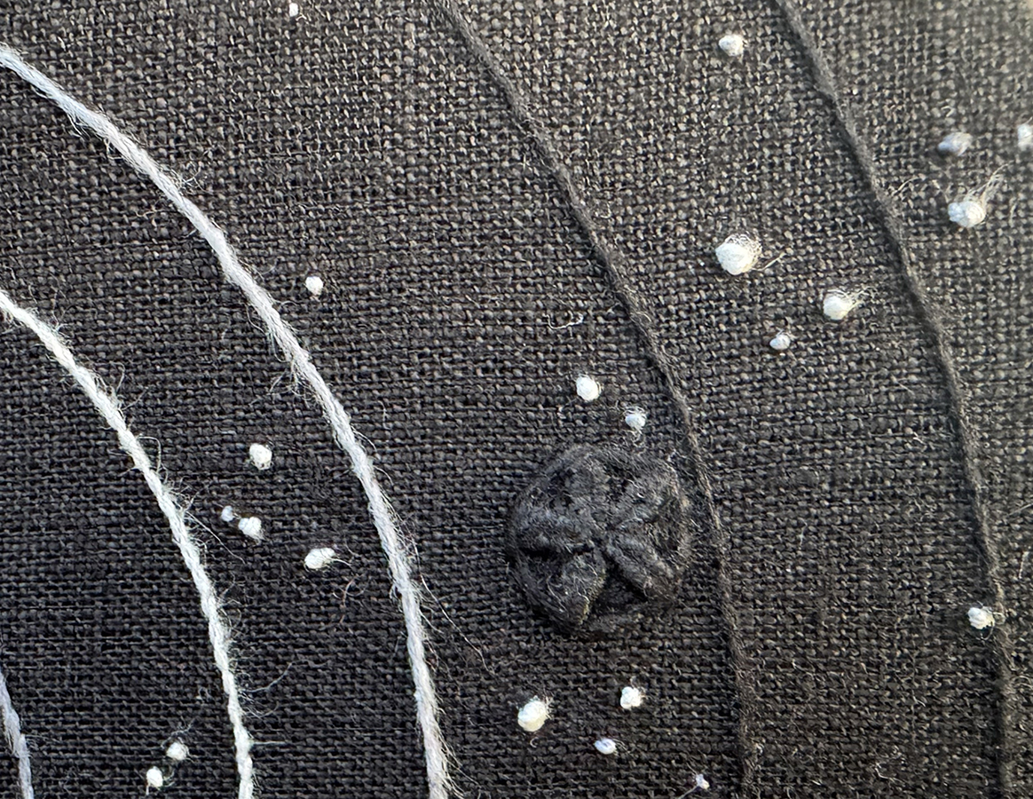 The embroidered Stranger on a background of orbits and small stars: it's black thread on a black linen evenweave background, but the texture makes it visible close up. The Stranger has 5 sets of 2 spines coming out from the center, which are done in whipped wheel stitches.