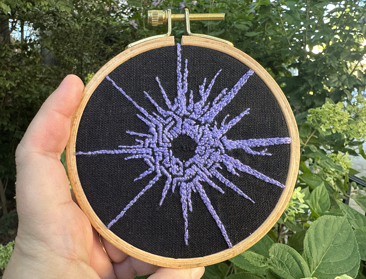 The symbol of the Eye of the Universe is embroidered in tight stitches of a slightly multitonal lavender linen thread on a black linen background that's placed in a small, wooden circular hoop. All the stitches are oriented towards the center of the Eye, so some sections are knobby while others look more like satin stitch. Most of the Eye's symbol has the angular nature of the Nomai's symbol for the Eye of the Universe, but the section between about 12 o'clock and 4 o'clock on an analog clock uses the branchier represenation of the Eye that the inhabitants of the Stranger use. In the center, the Eye's coordinates are stitched in a thin black thread that matches the black linen background.