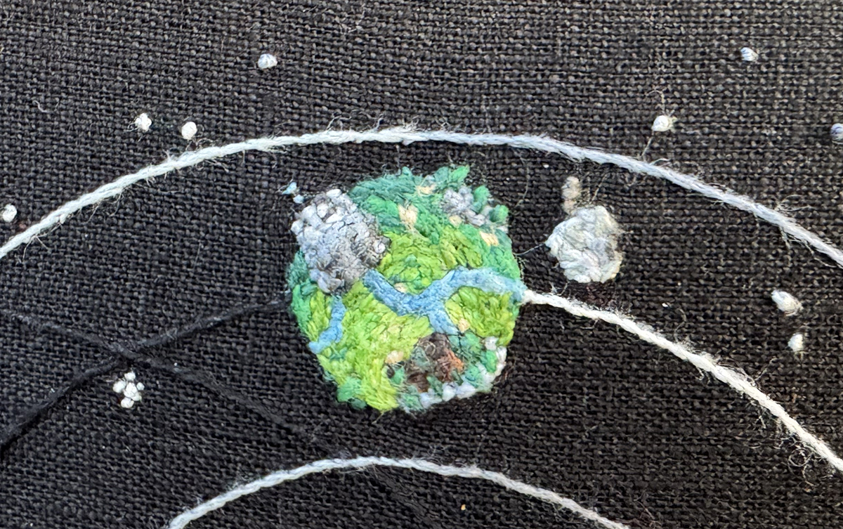 Timber Hearth in Outer Wilds solar system embroidery on a black linen background. Orbital paths for the Hourglass Twins, Timber Hearth, and Brittle Hollow can be seen in a gradient from white to black following the planets. Part of the Interloper's orbit can be seen. Timber Hearth is a gradient of greens in stem stitches from deep green at the top, farthest from the sun, to a bright lime at the bottom. Many trees in deeper greens dot the planet, as well as dirt patches in soft sand tones to deep browns in the settlement area. Blue rivers in chain stitch run down the planet. Rocky geysers are done in a variety of tight grey backstitch, and one is erupting with soft blue dots coming out into space. The Attlerock is a variety of tight grey stitches. It has the characteristic 'bite out of the apple' shape, and campfire smoke stitches can be seen floating away from the moon. Small French knots in whites and greys of varying thread thicknesses make up the starfield in the background.