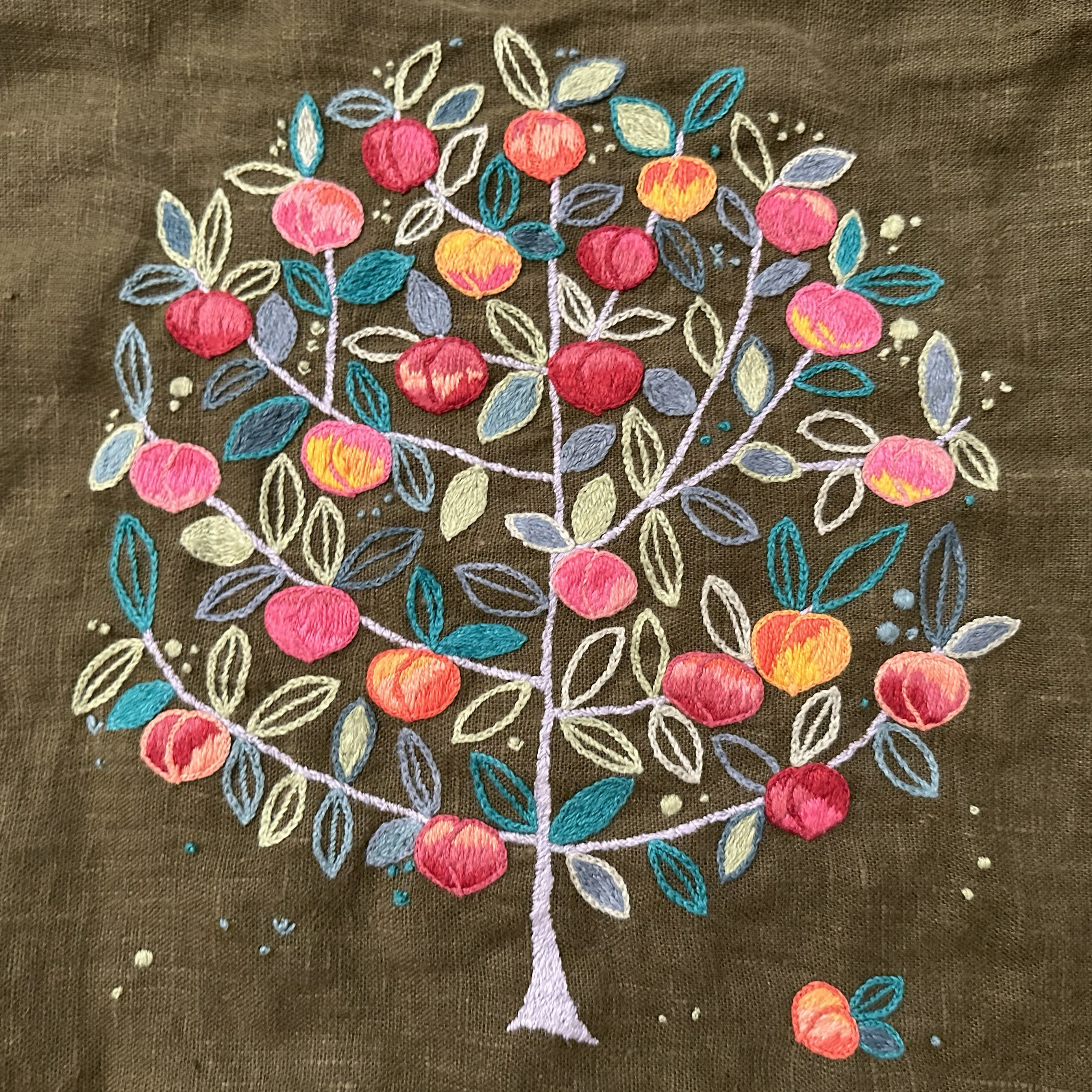 A peach tree is embroidered on a deep forest green linen background: the trunk and branches are made in a silver stem stitch that goes in lots of different directions to echo the feel of bark, there are simple leaves outlined in chain stitch in various greens and blues, some of the leaves are filled in satin stitch in complementary greens or blues, and there are peaches in all sorts of pinks, oranges, and reds. The peaches are also outlined in chain stitch, and they are all filled in colorblocked satin stiches in those pinks, oranges, and reds.