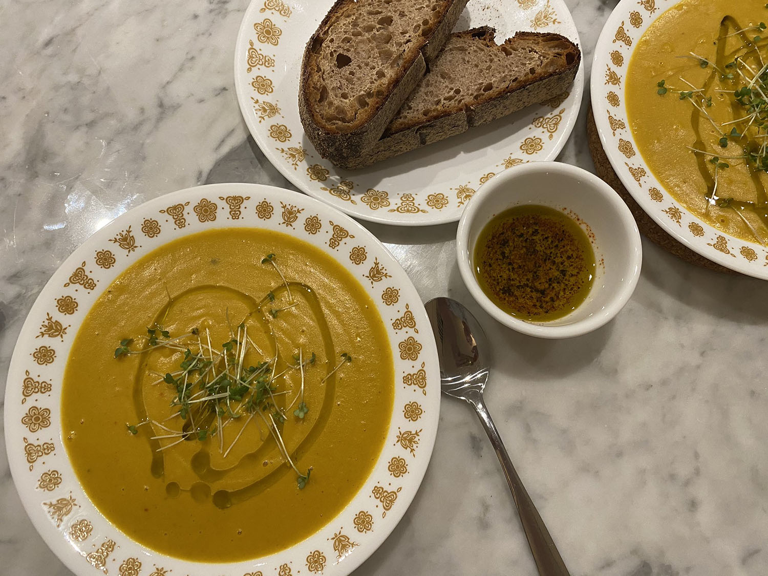 Photo of two shallow bowls (one half cut off) of a smooth roasted squash and red lentil soup topped with drizzled olive oil and mustard sprouts, a plate with two slices of a crusty bread, and a small bowl of olive oil with ground chilli and pepper for dipping the bread in.