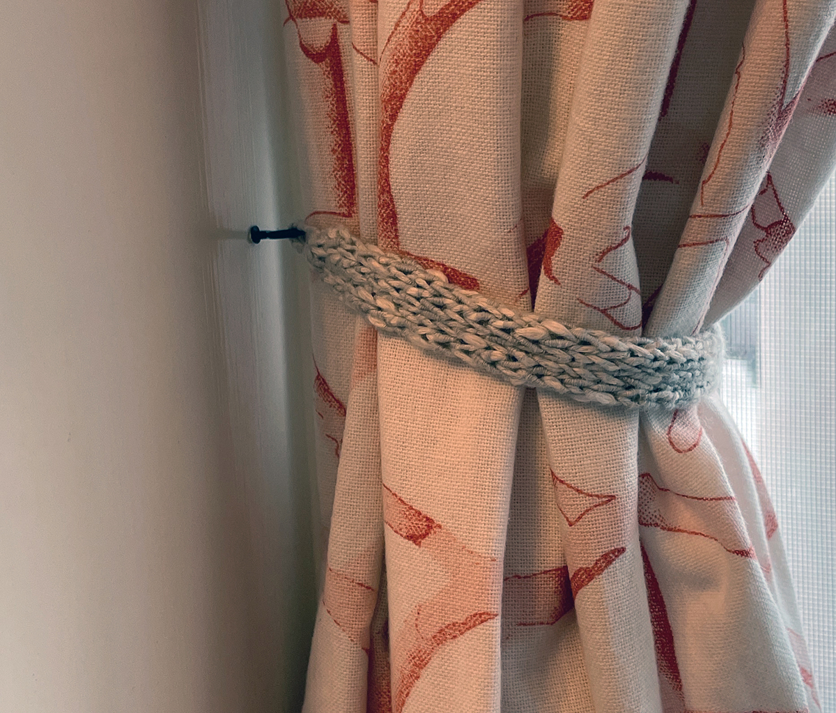 A cream colored curtain with a coral arches pattern is held back by a small knitted strip made of grey and white yarn that's looped onto a cup hook that's screwed into the inside of the window frame.