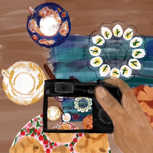 A hand holding a digital camera over a table set for a tea party: two teacups, jam, clotted cream, scones, deviled eggs, and a tart; the screen on the camera also shows the tea party spread