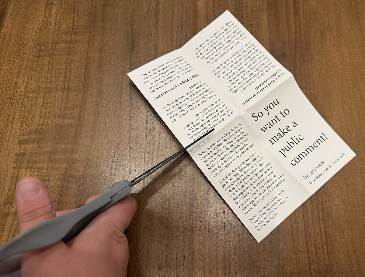 The mini-zine page is folded in half with short ends touching and a scissor is about to cut along the dotted line.