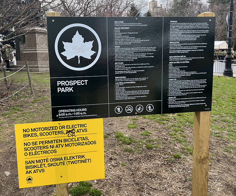 A yellow sign saying no motorized or electric bikes, scooters, and ATVs in three languages appended to Prospect Park's regular green rules sign