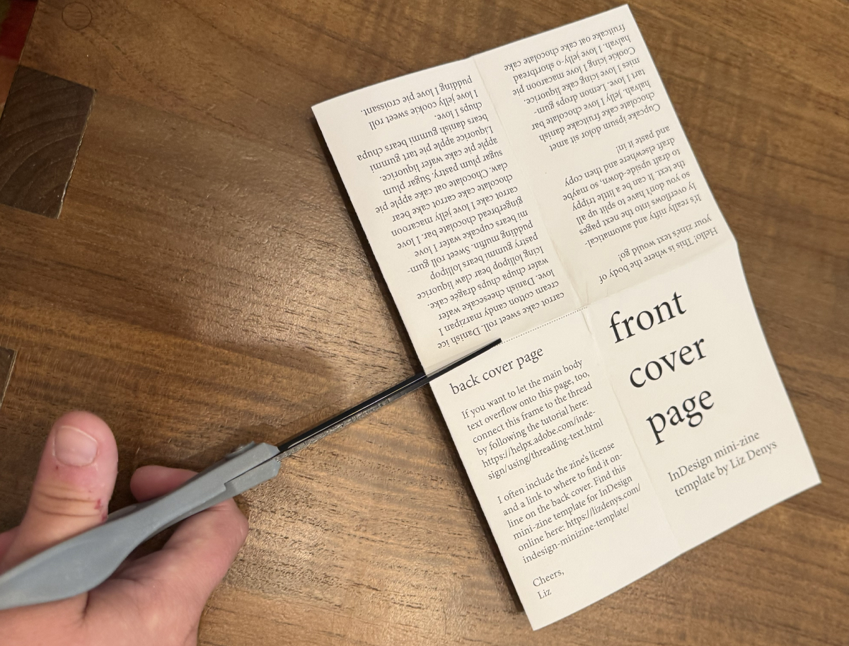 The mini-zine page is folded in half with short ends touching and a scissor is about to cut along the dotted line.